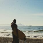 A lone surfer holding a surfboard looks out over the ocean waves at a sandy beach under a clear sky.
